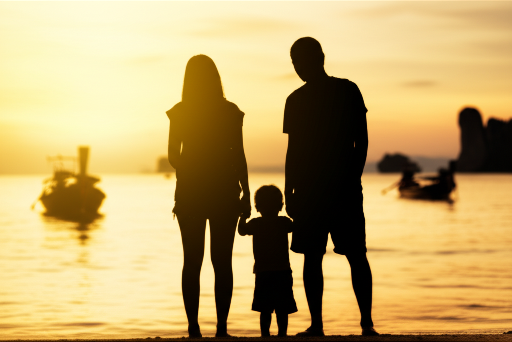 A gently blurred image showing a family (mother, father, young child) silhouetted against a warm, golden sunset on a pristine beach, with traditional longtail boats softly visible in the distance. The focus is on the feeling of tranquility and togetherness.